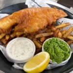 Delicious fish and chips served with tartar sauce, mushy peas, and a lemon wedge in a black dish.